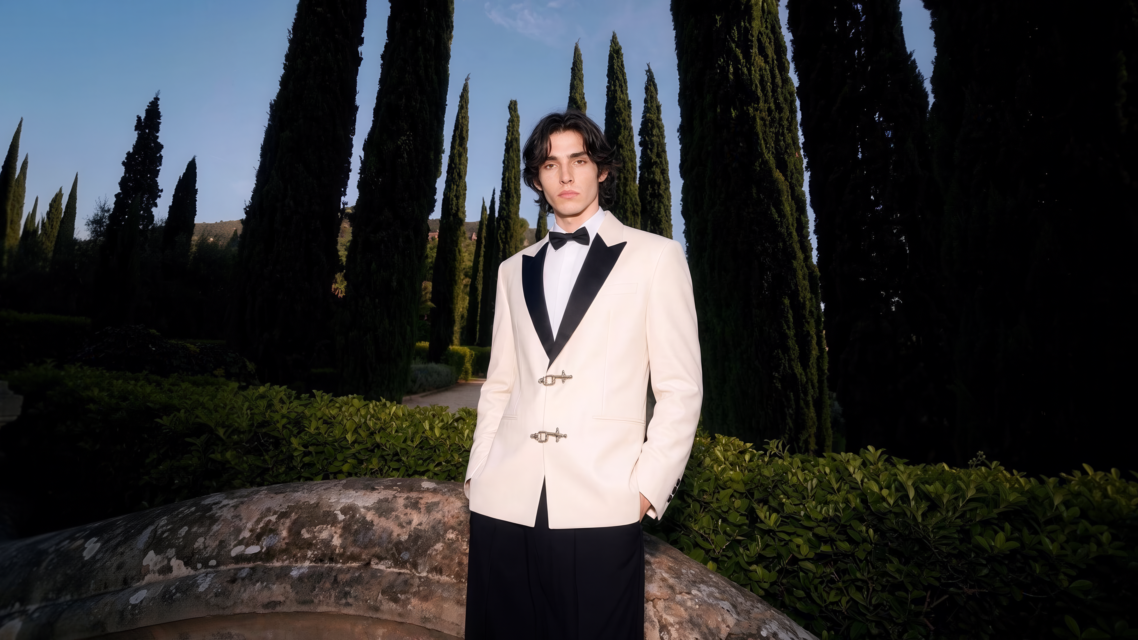 Man in white dinner jacket with black peak lapels standing before cypress trees in an evening garden